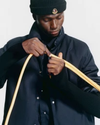 Black man wearing a dark beanie and puffer jacket holding yellow rope against a white background for Moncler Spring 2027.