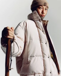 Model wearing a puffer jacket and knit beanie holding a wooden hiking stick against a plain background
