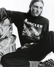 Moncler model wearing black graphic sweatshirt and dark pants sitting beside a stacked outdoor pack against a white background