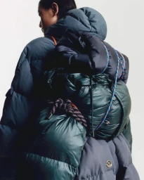 Model seen from behind wearing a puffer jacket and carrying a heavy loaded technical backpack against a white seamless background.