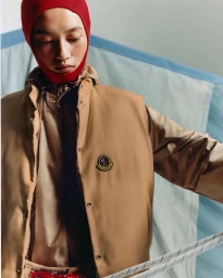 Model wearing a red balaclava and tan Moncler jacket against a light blue textured backdrop holding a rope.