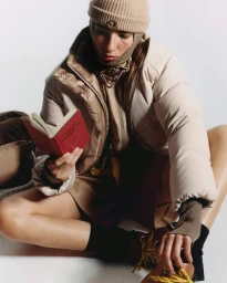 Model wearing a beige puffer jacket, beanie, and balaclava sitting on the floor while reading a small red book