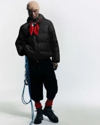Model wearing a black puffer jacket, red neck scarf, tan beanie, and climbing boots holding a blue rope on a white background.