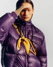 Model wearing a glossy purple puffer jacket with a yellow scarf against a plain studio background.