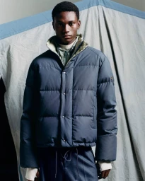 Young Black man wearing a navy blue quilted puffer jacket standing against a crinkled grey backdrop for Moncler.