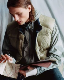 Model in olive padded vest and grey jacket holding a map and pen against a draped fabric backdrop