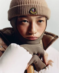 Close-up portrait of model wearing a brown knit beanie with a Moncler logo, balaclava, and puffer jacket.