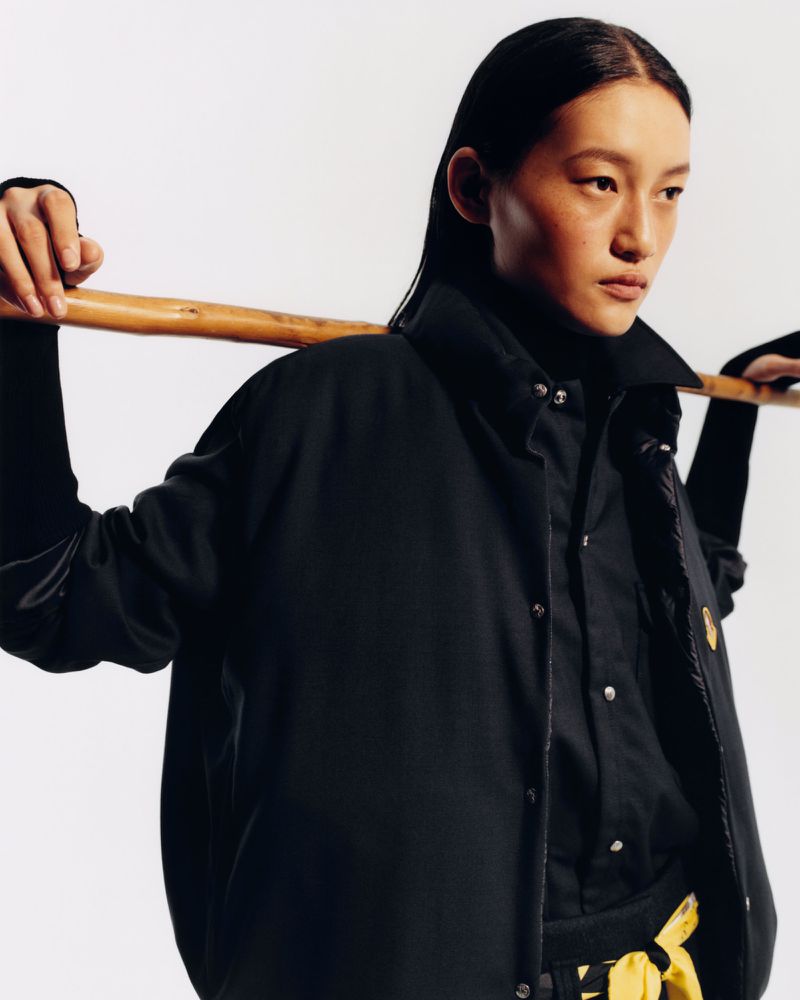 Model with dark hair wearing a black jacket holding a wooden stick across shoulders against a plain background