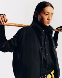 Model with dark hair wearing a black jacket holding a wooden stick across shoulders against a plain background