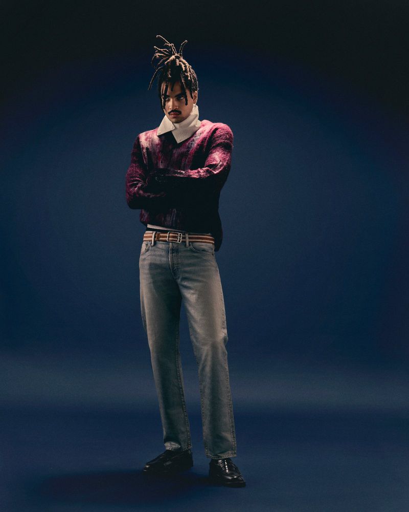 Full-length studio shot of an adult male wearing jeans, a sweater, and black dress shoes standing against a dark blue background.
