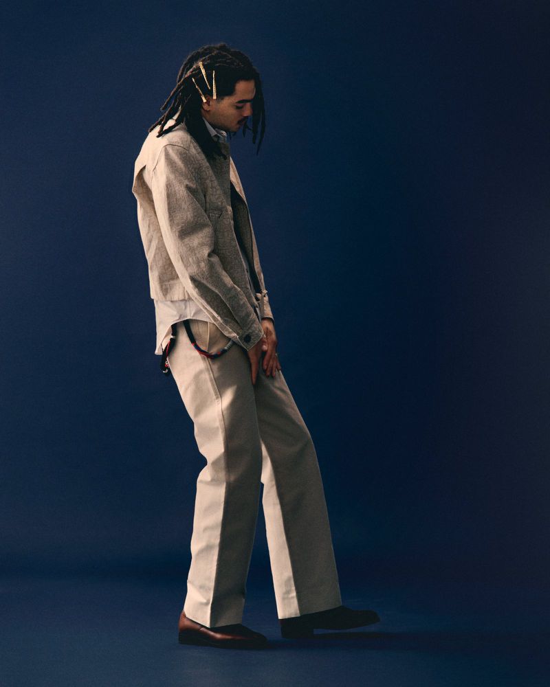 Studio shot of a young adult man with dreadlocks, wearing a neutral-toned jacket and pants, standing against a dark blue background.
