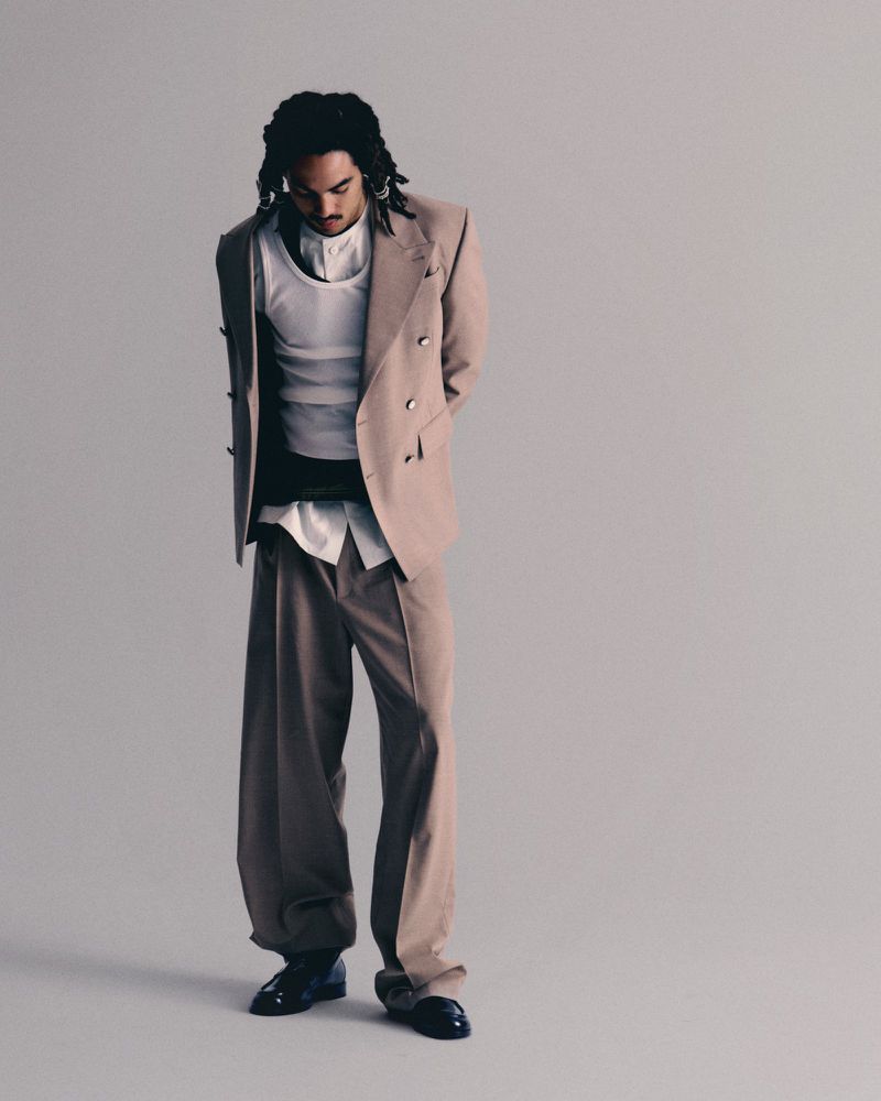 Full shot of a man wearing a tan suit and black shoes, standing against a light grey background in a studio setting with soft lighting.