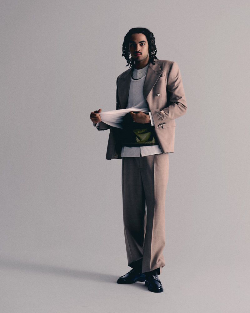 A stylish adult male stands in a photo studio wearing a tan suit, white shirt, and green bag, looking at a document he holds with both hands.