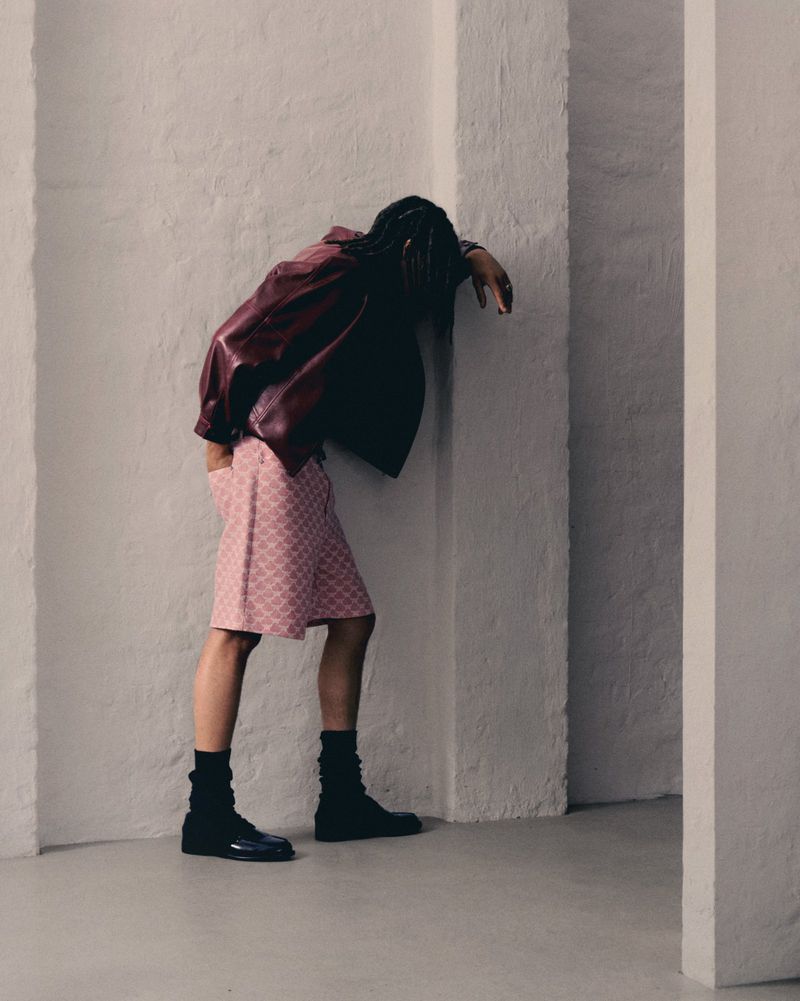 Fashion shot of adult male with dreadlocks in a burgundy jacket and pink shorts leaning against a white column in a minimalist setting.
