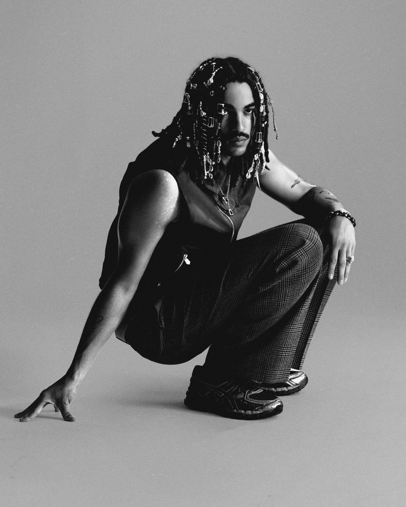 Portrait of a stylish adult man in a sleeveless top and plaid pants, crouching indoors in a photo studio against a grey backdrop.