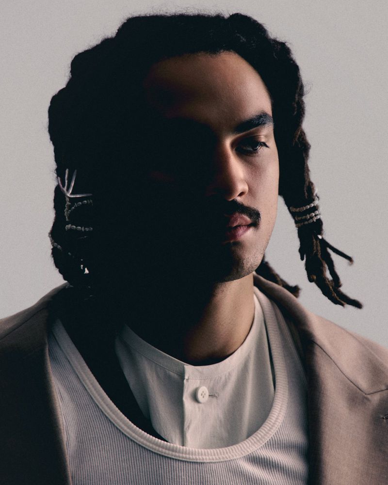 Portrait of a man with dreadlocks wearing a white undershirt and a beige blazer in a studio with soft, diffused lighting.