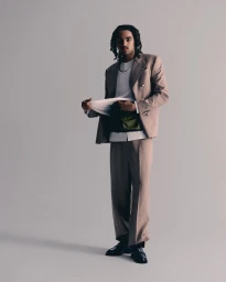 Musician Badchieff posing in a tailored beige double-breasted suit and black dress shoes in front of a neutral grey studio backdrop.