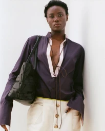 Model Janet Jumbo wearing a dark purple tunic and holding a black woven handbag against a white studio wall