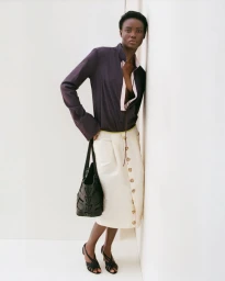 Model Janet Jumbo leaning against a white studio corner wearing a dark blouse and cream skirt holding a black handbag