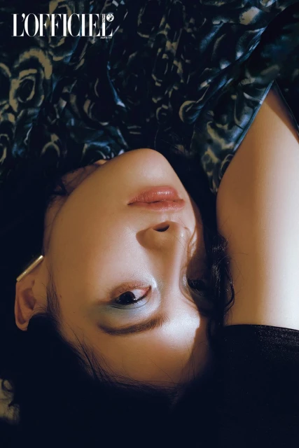 Close-up of a woman with blue eyeshadow and gold earrings looking up, with dark floral-patterned fabric in the background, editorial.