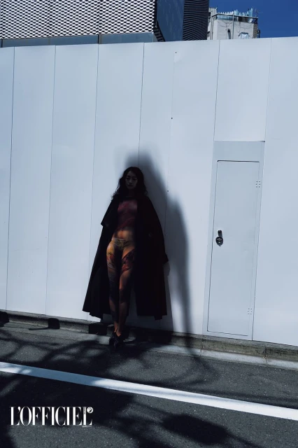 Full-length shot of an adult female model wearing a coat and patterned tights, posing against a white wall in an urban environment.