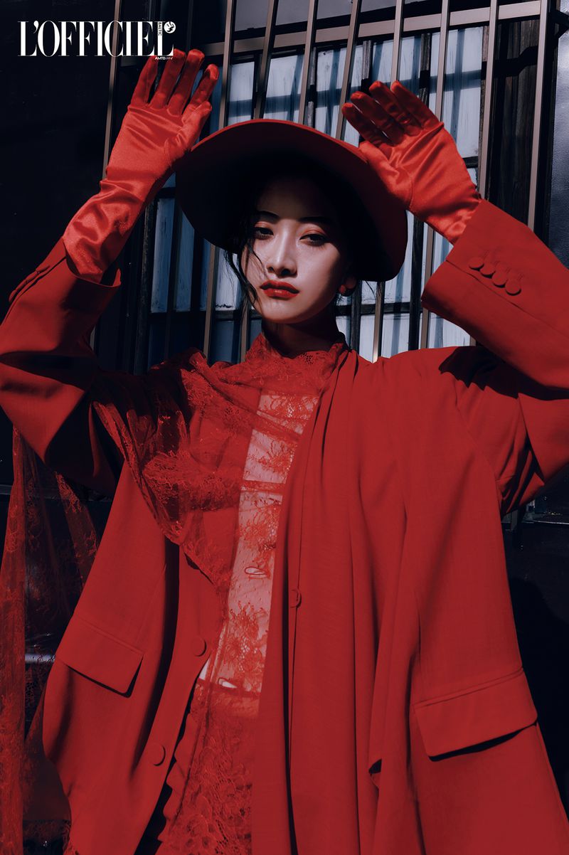 Portrait of a woman in a red outfit with a hat and gloves, posing in front of a metal gate with the 'L'Officiel' logo visible.