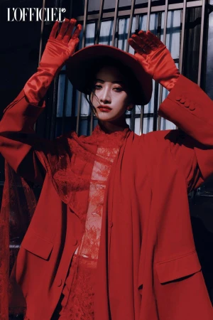 Portrait of a woman in a red outfit with a hat and gloves, posing in front of a metal gate with the 'L'Officiel' logo visible.