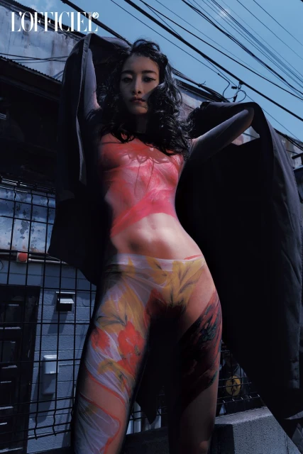 A woman with body paint poses outside, wearing a dark jacket with a city street, buildings, and an overhead power line as a backdrop.