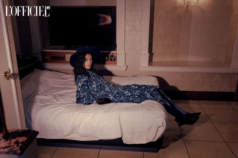 Editorial fashion shot of a woman in a patterned dress and hat lying on a bed in a hotel room with neutral wallpaper and tile.