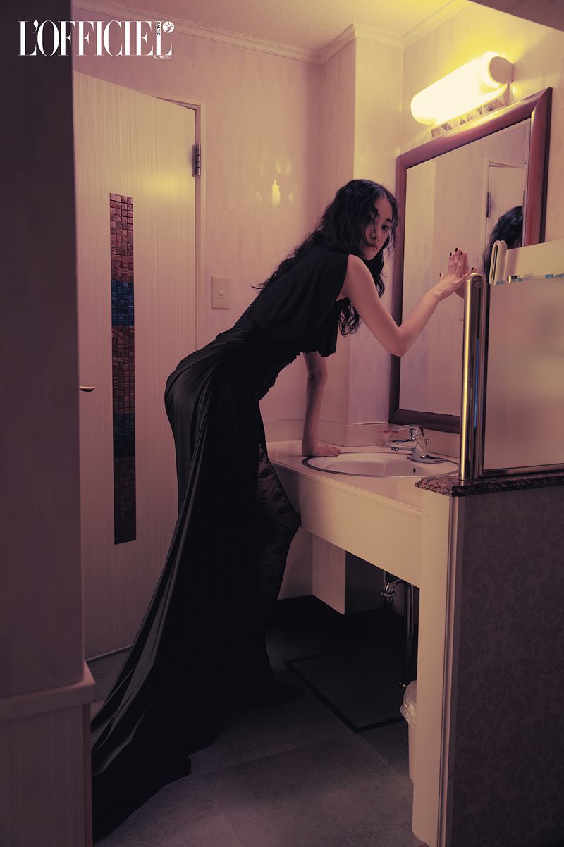 A woman in a long black dress poses in a bathroom with a vintage vibe. The soft lighting enhances the overall aesthetic.