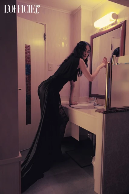 A woman in a long black dress poses in a bathroom with a vintage vibe. The soft lighting enhances the overall aesthetic.