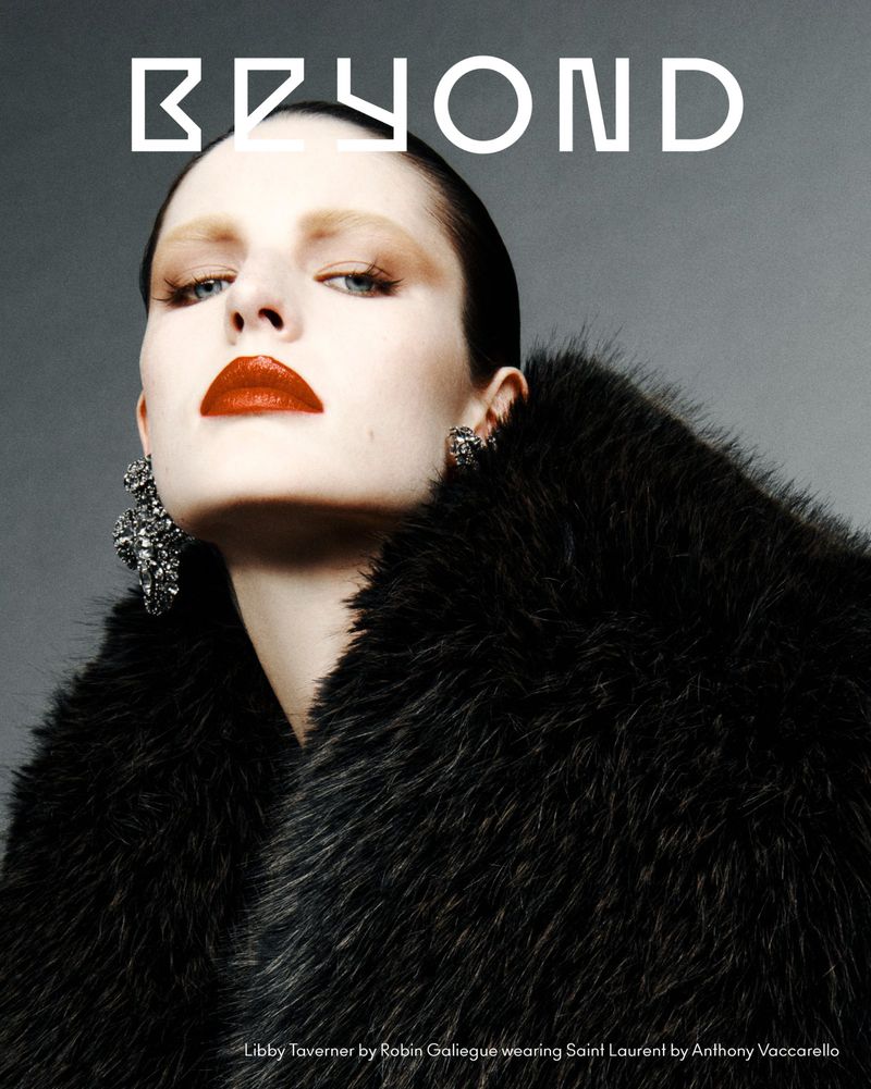 Model with slicked-back dark hair and red lipstick wearing a black faux-fur coat against a grey studio backdrop.