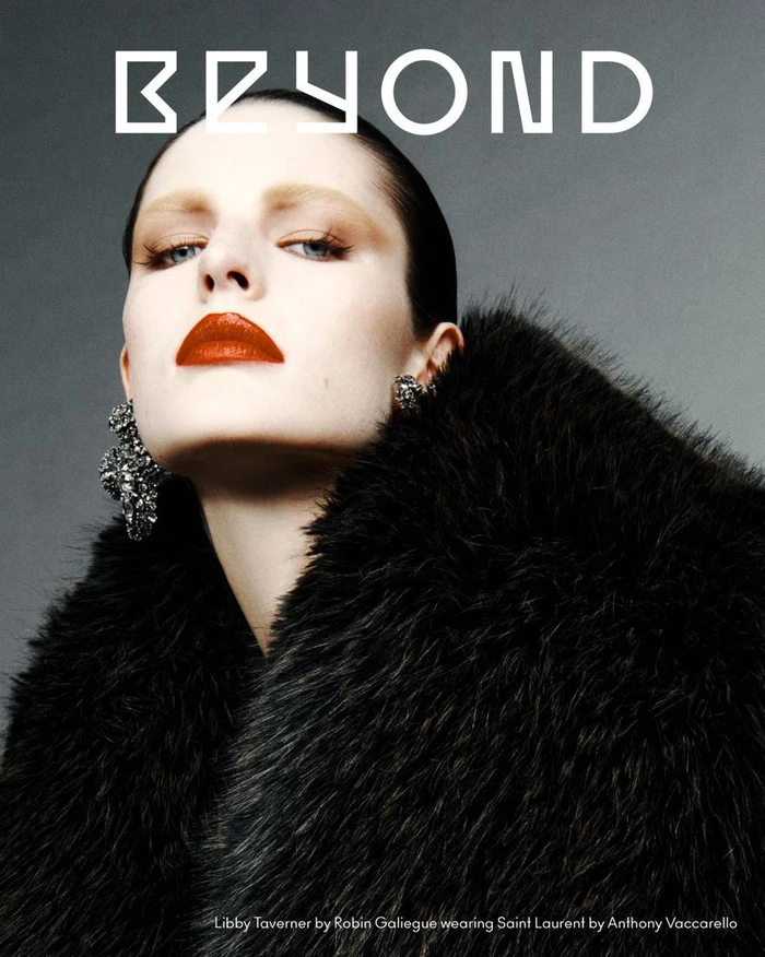 Model with slicked-back dark hair and red lipstick wearing a black faux-fur coat against a grey studio backdrop.