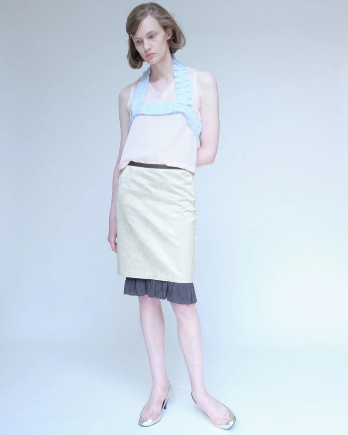 Model wearing a pale pink top with a light blue ruffled harness and beige skirt against a neutral background