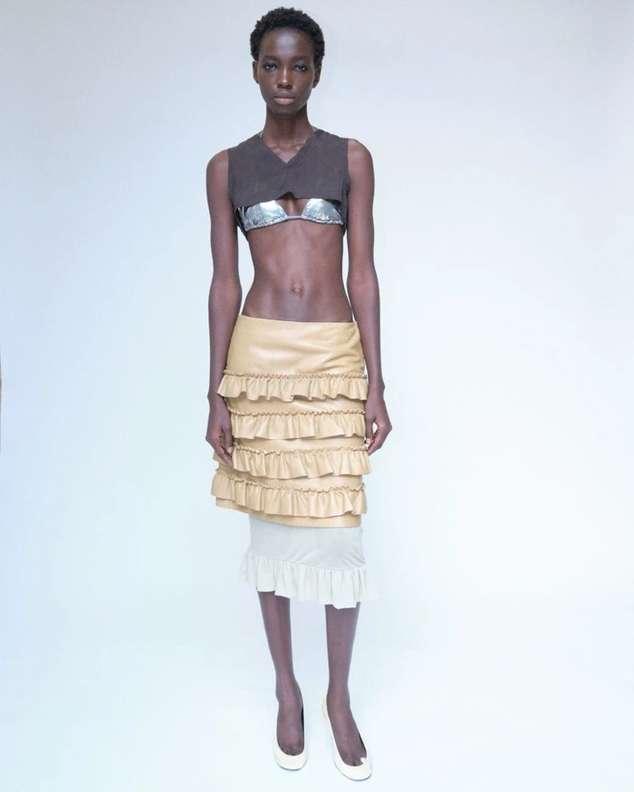 Model standing against a white seamless backdrop wearing a brown crop top, layered ruffled beige skirt, and white shoes.