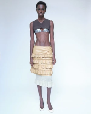 Model standing against a white seamless backdrop wearing a brown crop top, layered ruffled beige skirt, and white shoes.