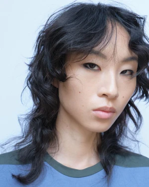Close-up portrait of a model with dark curly layered hair wearing a blue and green top against a white background for Paloma Wool.