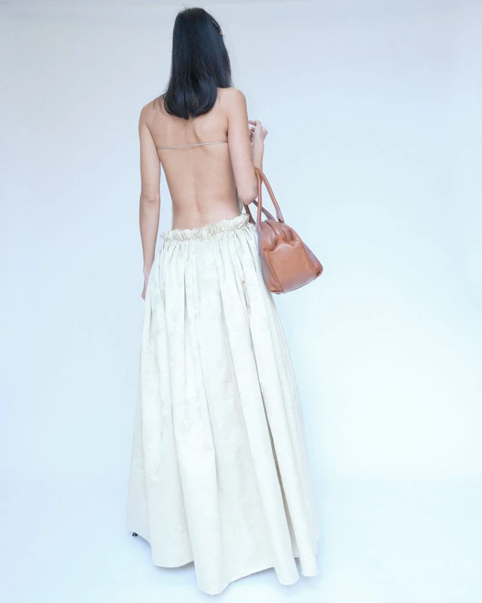 Model with long dark hair seen from behind wearing an off-the-shoulder cream skirt and holding a tan leather handbag.