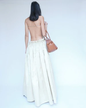 Model with long dark hair seen from behind wearing an off-the-shoulder cream skirt and holding a tan leather handbag.