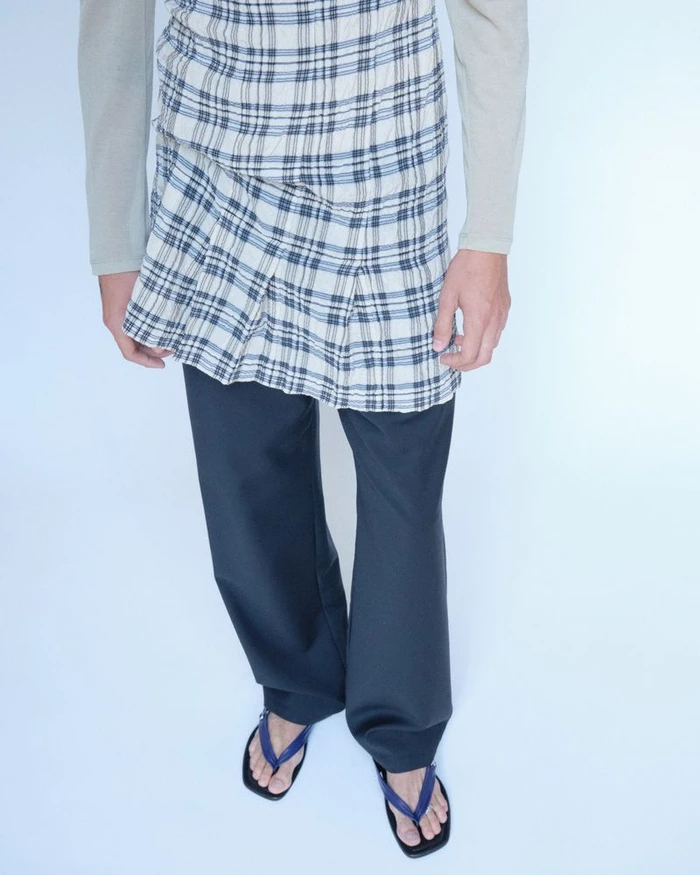 Model wearing a plaid wrap skirt over dark trousers and thong sandals against a pale background