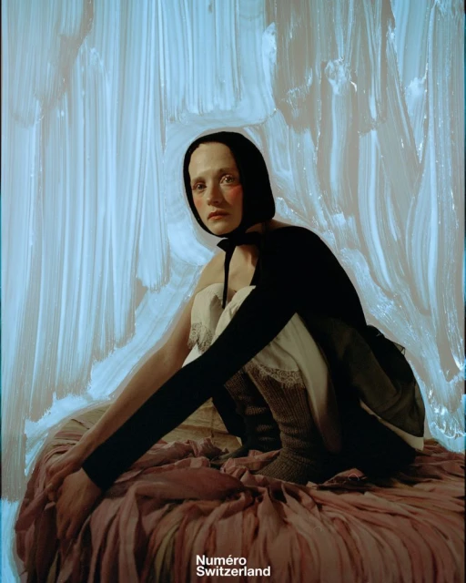 A pale, red-cheeked fashion model poses indoors against an abstract painted backdrop, wearing a black bonnet, long black sleeves, and layered fabrics.