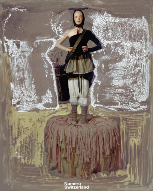 Fashion editorial shot of a pale-skinned woman in avant-garde apparel posing on a draped pedestal in front of a textured neutral backdrop.