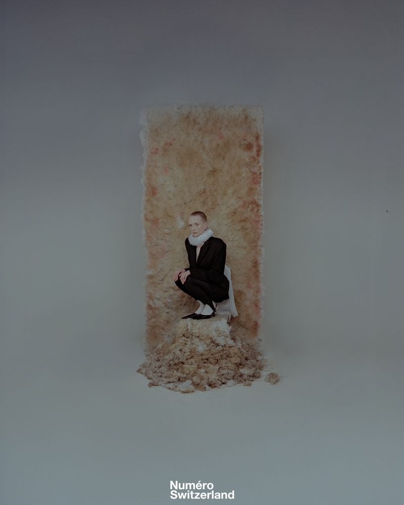 Fashion editorial shot of a woman in a dark suit crouching against a tan-colored backdrop with the magazine logo at the bottom.