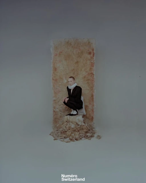 Fashion editorial shot of a woman in a dark suit crouching against a tan-colored backdrop with the magazine logo at the bottom.