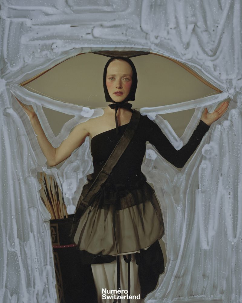 Artistic portrait of a fair-skinned woman in a black dress and head covering, posing in front of a textured abstract background.