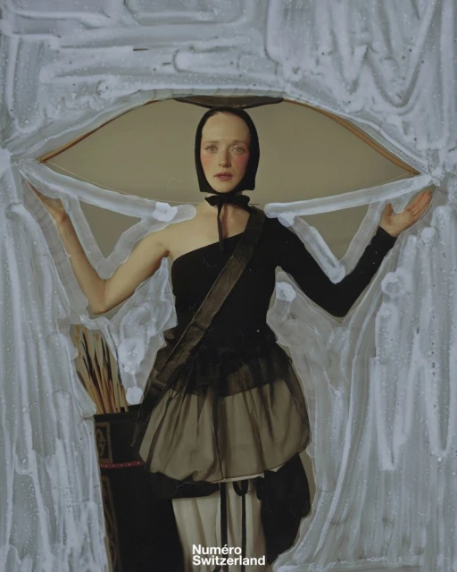 Artistic portrait of a fair-skinned woman in a black dress and head covering, posing in front of a textured abstract background.