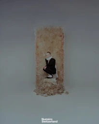 Model Olga Zapivokhina crouching in a black suit with a white ruff collar on a textured pedestal against a neutral studio backdrop.
