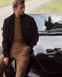 Man wearing a black jacket and brown trousers leaning against a black car for a BOSS campaign by James Yates.