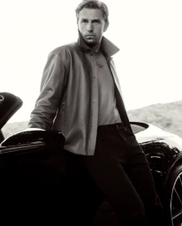 Man leaning against a sleek sports car wearing a leather jacket and dark trousers in an outdoor setting.
