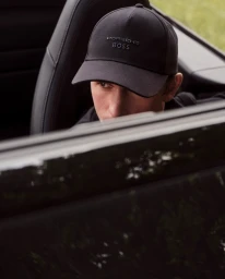 Man wearing a black Porsche BOSS baseball cap seated in a convertible car with dark leather seats.